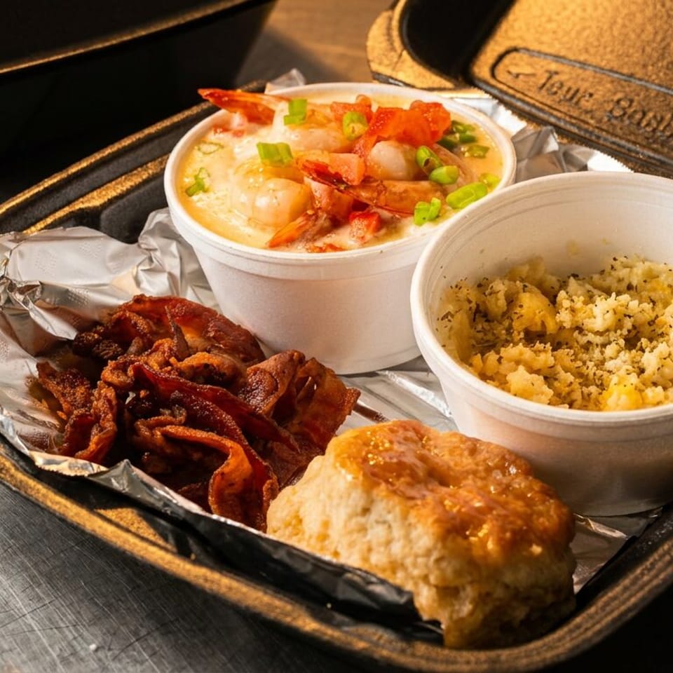 Takeout meal featuring creamy shrimp topped with bacon and green onions, seasoned rice, crispy bacon, and a golden biscuit.