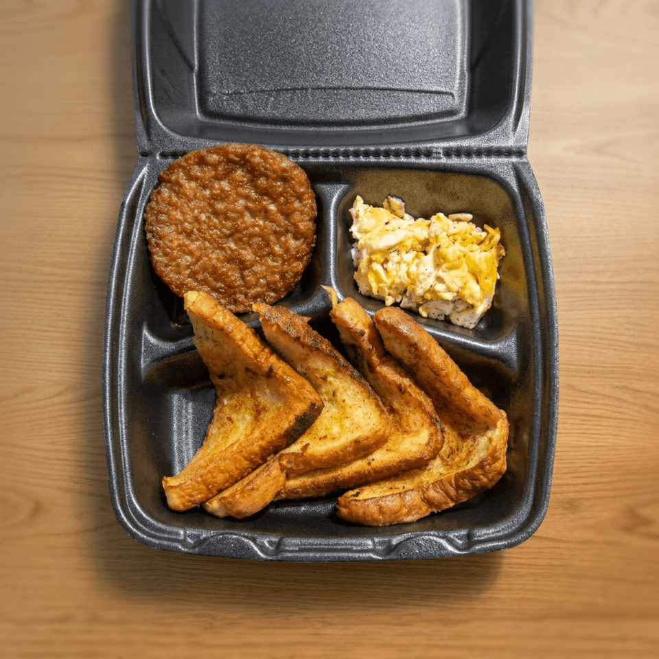Black takeout container holding French toast triangles, a sausage patty, and scrambled eggs on a wooden table.